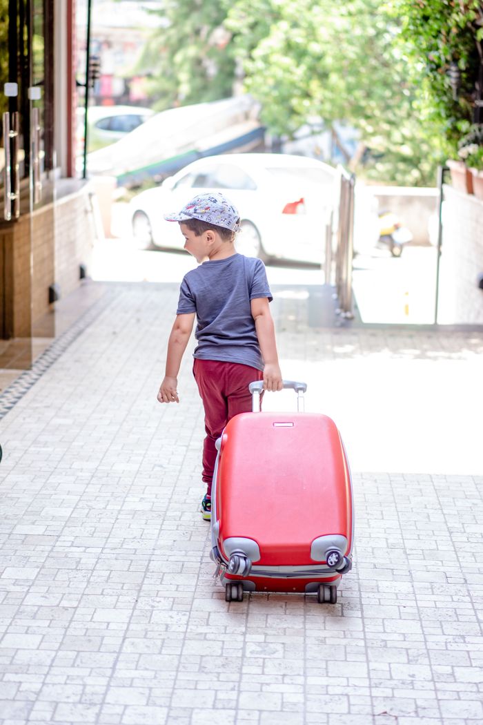 A young boy in a gray shirt and red pants pulling a suitcase outdoors.
